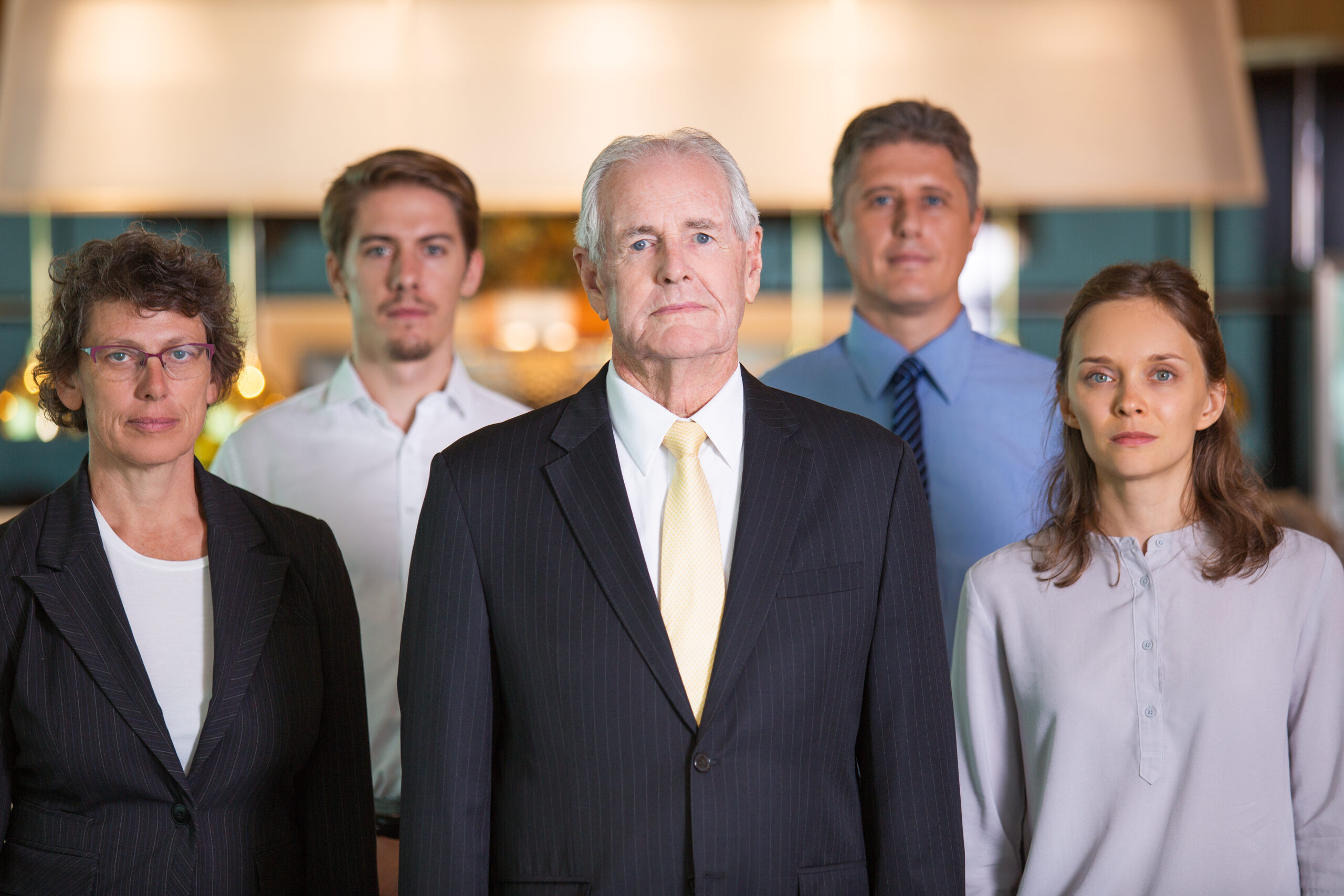 Closeup of business team of five serious diverse people standing and looking at camera with blurred view in background. Two blurred businessmen are in background. Front view.